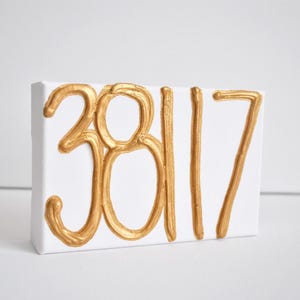 Może przedstawiać: Białe płótno z namalowanymi złotymi cyframi 38117. Grube, teksturowane cyfry kontrastują z czystym, białym tłem. Prostokątne płótno jest naciągnięte na drewnianą ramę, tworząc prosty, nowoczesny wygląd.