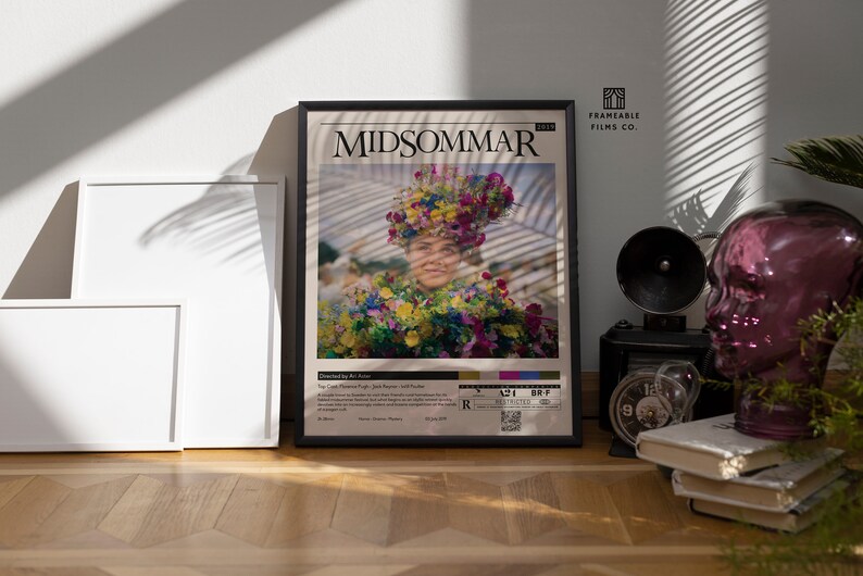 May include: A framed movie poster for the film "Midsommar" featuring a woman wearing a flower crown and a floral dress. The poster is black framed and has a white background with the movie title in black text. The poster also includes the movie's rating, runtime, and release date.