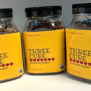 May include: Three clear plastic jars filled with dark brown coconut sugar cubes. Each jar features a yellow label with the text "THREE CUBE coconut sugar" and a row of red hearts. The labels also state "BY AROMA VAN JAVA" and "8.83 OZ (250 GR)".