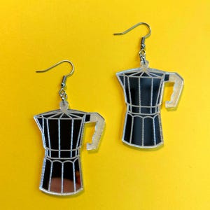 May include: A pair of coffee pot-shaped earrings. The earrings are black and silver, with a detailed outline of a stovetop espresso maker. The earrings hang from silver-colored fish hook ear wires. The background is a solid yellow.
