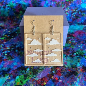 May include: Clear acrylic earrings with a rectangular design. Each earring features a cloud and star motif with the words "Moment Land" in purple. The earrings hang from silver-colored fish hook ear wires. The earrings are displayed on a brown card.