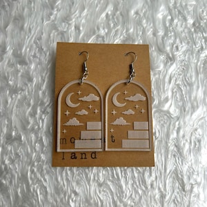 May include: A pair of clear acrylic earrings with a celestial design. Each earring features a crescent moon, clouds, stars, and a staircase within an arched frame. The word "moont land" is printed at the bottom of the earrings.
