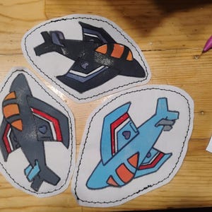 May include: Three embroidered patches featuring stylized airplane designs. One patch is dark gray with orange accents, another is medium gray with red and orange details, and the third is light blue with red and orange highlights, all with black stitching.