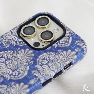 May include: A blue and white patterned phone case with gold camera lens protectors. The case features a detailed floral design and a black border around the camera lenses. The lens protectors are adorned with small, clear crystals.