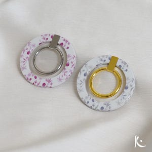 May include: Two floral phone ring holders. One has a silver ring and holder, the other has a gold ring and holder. Both have a white background with a floral pattern in shades of purple and grey.