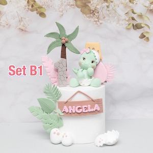 Puede incluir: Una tarta blanca decorada con un tema de dinosaurio beb&eacute;. La tarta presenta un dinosaurio verde, una palmera, el n&uacute;mero uno y el nombre "Angela" en un letrero de madera. Hojas rosas y verdes y c&aacute;scaras de huevo completan el dise&ntilde;o.