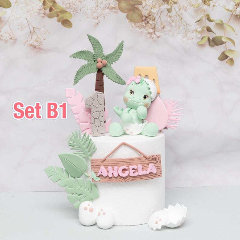 Puede incluir: Una tarta blanca decorada con un tema de dinosaurio beb&eacute;. La tarta presenta un dinosaurio verde, una palmera, el n&uacute;mero uno y el nombre "Angela" en un letrero de madera. Hojas rosas y verdes y c&aacute;scaras de huevo completan el dise&ntilde;o.