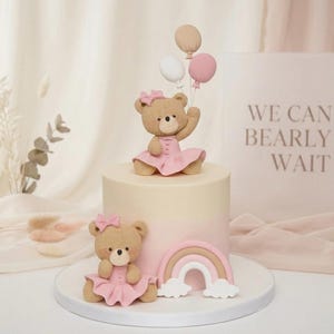 May include: A two-tier cake featuring two tan teddy bears wearing pink dresses and bows. The top bear holds three balloons in white, tan, and pink. The bottom bear sits next to a small rainbow with white clouds. The cake is light pink and cream.