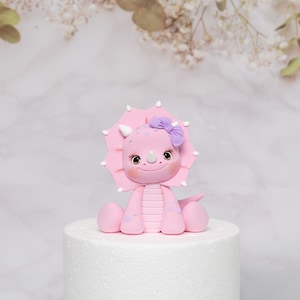 Puede incluir: Figura de triceratops rosa con un lazo morado en la cabeza. El dinosaurio tiene una cara sonriente, cuernos blancos y est&aacute; sentado sobre una base blanca para pastel. Decoraci&oacute;n.