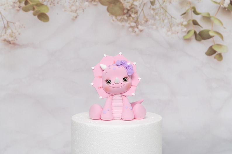 Puede incluir: Figura de triceratops rosa con un lazo morado en la cabeza. El dinosaurio tiene una cara sonriente, cuernos blancos y est&aacute; sentado sobre una base blanca para pastel. Decoraci&oacute;n.