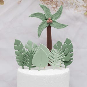 Puede incluir: Decoraci&oacute;n para tarta con una palmera y hojas tropicales. La palmera tiene un tronco marr&oacute;n, hojas verdes y tres peque&ntilde;os cocos rosas. Las hojas son de varios tonos de verde, creando un tema de jungla.