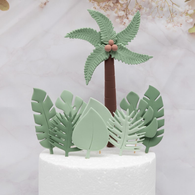 Puede incluir: Decoraci&oacute;n para tarta con una palmera y hojas tropicales. La palmera tiene un tronco marr&oacute;n, hojas verdes y tres peque&ntilde;os cocos rosas. Las hojas son de varios tonos de verde, creando un tema de jungla.