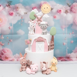 May include: A two-tiered cake decorated with a pink barn, a windmill, a sun, clouds, and farm animals. The animals include a goat, a sheep, a rabbit, a pig, a horse, and a chick. The cake is decorated with a pink and white color scheme.