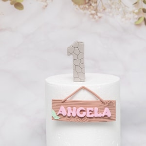 Puede incluir: Un adorno para tarta con el n&uacute;mero uno gris con un patr&oacute;n texturizado y un letrero de madera con el nombre "ANGELA" en rosa. El letrero tiene un detalle de hoja y est&aacute; suspendido por una cuerda rosa.
