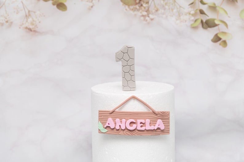 Puede incluir: Un adorno para tarta con el n&uacute;mero uno gris con un patr&oacute;n texturizado y un letrero de madera con el nombre "ANGELA" en rosa. El letrero tiene un detalle de hoja y est&aacute; suspendido por una cuerda rosa.