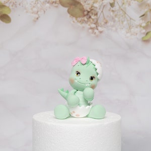 Puede incluir: Una figura de dinosaurio verde menta con un lazo rosa, sentada sobre una base blanca. El dinosaurio tiene una cara sonriente, mejillas rosadas y lleva un pa&ntilde;al. El fondo es un patr&oacute;n de m&aacute;rmol con acentos florales.