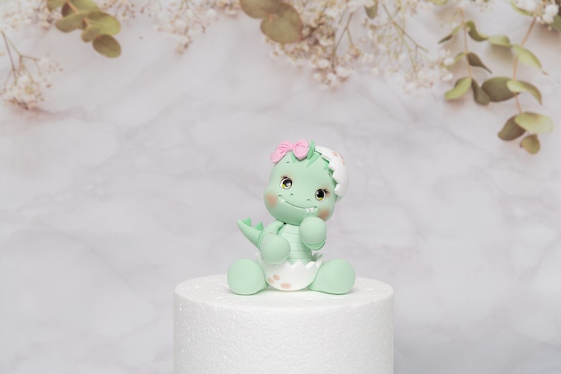 Puede incluir: Una figura de dinosaurio verde menta con un lazo rosa, sentada sobre una base blanca. El dinosaurio tiene una cara sonriente, mejillas rosadas y lleva un pa&ntilde;al. El fondo es un patr&oacute;n de m&aacute;rmol con acentos florales.