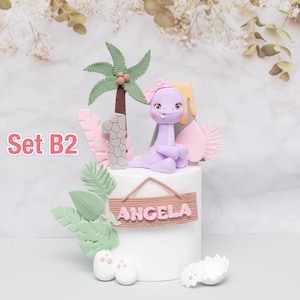 Puede incluir: Una tarta blanca decorada con un dinosaurio morado, una palmera y el nombre "Angela" en un letrero de madera. La tarta tambi&eacute;n presenta el n&uacute;mero 1, detalles en rosa y el texto "Set B2".