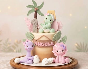 Op de afbeelding: Een versierde taart met dinosaurusfiguren, een palmboom en de naam "Angela". De taart heeft een groene dinosaurus die uit een ei komt, een paarse dinosaurus en een roze triceratops. De taart staat op een houten serveerplank.
