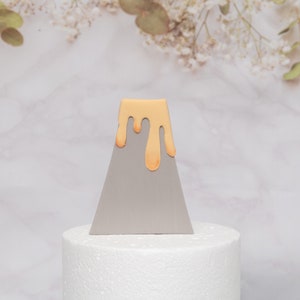 Puede incluir: Un adorno para tarta triangular gris con un dise&ntilde;o de goteo naranja claro. El adorno para tarta se muestra sobre una base redonda blanca, con un fondo de m&aacute;rmol y detalles florales.