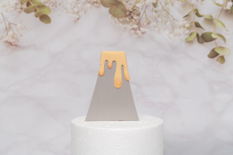 Puede incluir: Un adorno para tarta triangular gris con un dise&ntilde;o de goteo naranja claro. El adorno para tarta se muestra sobre una base redonda blanca, con un fondo de m&aacute;rmol y detalles florales.