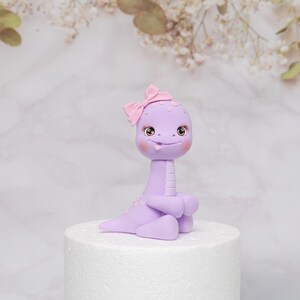Puede incluir: Una caprichosa figura de dinosaurio de color lavanda con un lazo rosa en la cabeza. La figura tiene ojos grandes y expresivos, mejillas rosadas y una peque&ntilde;a lengua. Est&aacute; sobre una base redonda blanca.