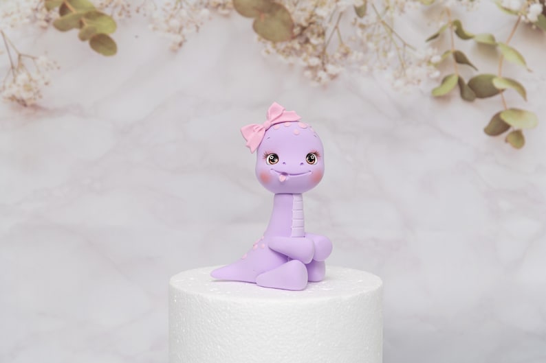 Puede incluir: Una caprichosa figura de dinosaurio de color lavanda con un lazo rosa en la cabeza. La figura tiene ojos grandes y expresivos, mejillas rosadas y una peque&ntilde;a lengua. Est&aacute; sobre una base redonda blanca.