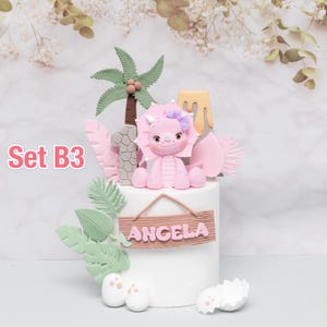 Puede incluir: Adorno para tarta con un triceratops rosa, una palmera y el n&uacute;mero 1. La tarta est&aacute; decorada con hojas verdes, detalles rosas y un letrero de madera con la palabra "ANGELA". Tambi&eacute;n se ve el texto "Set B3".