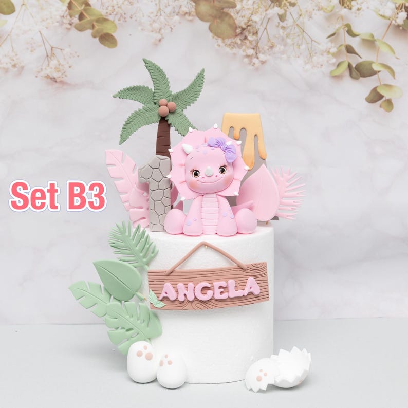 Puede incluir: Adorno para tarta con un triceratops rosa, una palmera y el n&uacute;mero 1. La tarta est&aacute; decorada con hojas verdes, detalles rosas y un letrero de madera con la palabra "ANGELA". Tambi&eacute;n se ve el texto "Set B3".