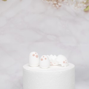 Puede incluir: Adornos para tartas de huevos de Pascua blancos con lunares rosas y una c&aacute;scara de huevo agrietada. Los adornos est&aacute;n sobre un soporte blanco para tartas, con un fondo de m&aacute;rmol y ramitas de vegetaci&oacute;n.