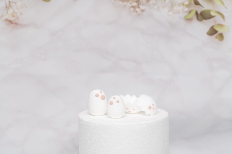 Puede incluir: Adornos para tartas de huevos de Pascua blancos con lunares rosas y una c&aacute;scara de huevo agrietada. Los adornos est&aacute;n sobre un soporte blanco para tartas, con un fondo de m&aacute;rmol y ramitas de vegetaci&oacute;n.