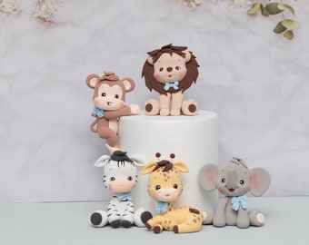 Op de afbeelding: Vijf schattige dierlijke taarttoppers gemaakt van fondant. De toppers omvatten een leeuw, een aap, een zebra, een giraffe en een olifant. Elk dier draagt ​​een blauwe strik en heeft een vriendelijke uitdrukking.