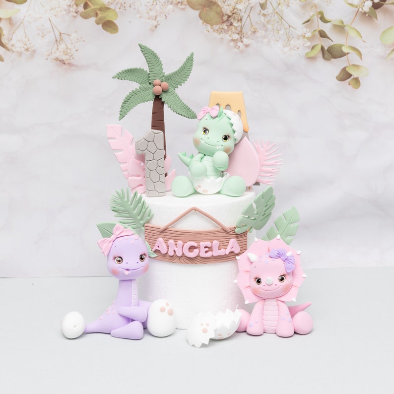 Puede incluir: Un adorno para tarta caprichoso con una base blanca adornada con figuras de dinosaurios en colores pastel, palmeras y hojas decorativas. La tarta est&aacute; personalizada con el nombre "Angela" y el n&uacute;mero "1".