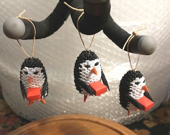 May include: Three origami penguin ornaments. Each penguin is black and white with a red beak and feet. They are hanging from a black bar with gray spheres on each end, suspended by twine.