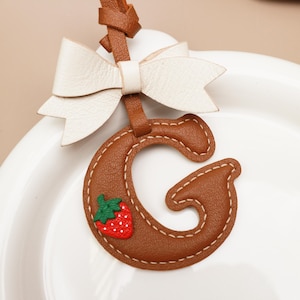 Op de afbeelding: Een bruinleren letter "G" sleutelhanger met een witte strik en een kleine rode aardbeienapplicatie. De letter is gestikt met wit garen en heeft een leren riem. De strik is gemaakt van wit leer.