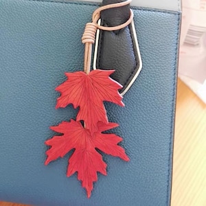 Könnte beinhalten: Ein roter Ahornblatt-Anhänger hängt an einem Lederriemen, der an einer blauen Handtasche befestigt ist. Der Anhänger zeigt zwei detaillierte Blätter mit filigranen Adern. Die Tasche hat einen schwarzen Griff mit weißer Umrandung.