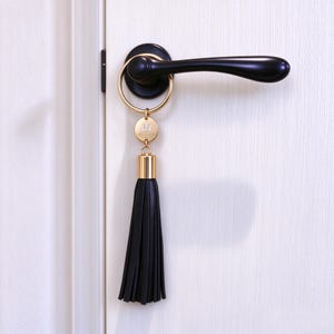 May include: A black leather tassel keyring featuring a gold-toned circular charm with an engraved emblem, attached to a gold-toned metal ring. The keyring is hanging from a black door handle mounted on a white door with a visible vertical frame.