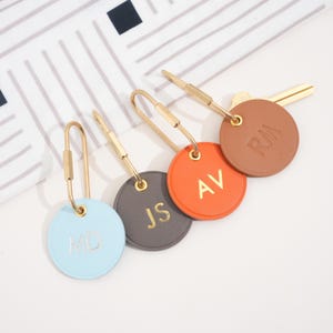 May include: Four personalized keychains in various colors: light blue, gray, orange, and brown. Each keychain features a gold-colored clasp and a circular tag with gold initials. The initials are "MD", "JS", "AV", and "RM".