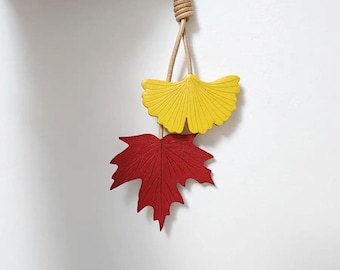 May include: A decorative hanging ornament featuring a yellow ginkgo leaf and a red maple leaf. The leaves are suspended from a natural-colored rope, creating a fall-themed accent. The leaves are painted with a matte finish.