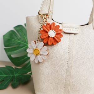 Può includere: Una borsa a mano color crema con chiusura dorata e portachiavi a forma di fiore di margherita. Il portachiavi presenta due ciondoli a forma di margherita, uno arancione e uno bianco, con centri gialli e bianchi. La borsa ha il testo "LOUIS SILMA".