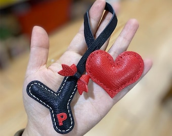 Puede incluir: Un llavero de cuero negro con un dije de corazón de cuero rojo. El llavero tiene una letra "P" cosida en él.