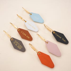 May include: A selection of personalised keyrings in shades of blue, white, brown, grey, pink, orange, and black. Each keyring has a gold-coloured metal ring and is engraved with custom text, including names and dates.
