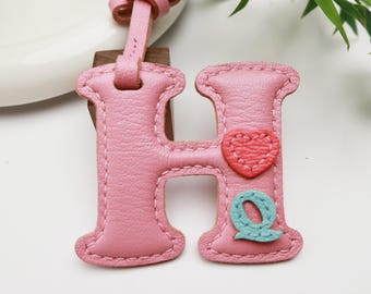Puede incluir: Llavero de cuero rosa con la letra "H", un corazón rojo y una "Q" turquesa. El llavero tiene un lazo rosa y una correa de cuero para colgarlo. Es probable que sea un accesorio personalizado.