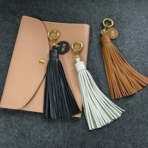 Puede incluir: Tres llaveros con borlas de cuero en negro, blanco y marrón, cada uno con un anillo dorado y una pequeña etiqueta redonda con iniciales. Una cartera de cuero color rubor en el fondo.