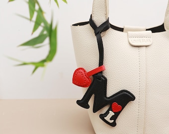 Puede incluir: Bolso de mano color crema con detalles en negro y un charm decorativo. El charm presenta un corazón rojo y las letras "M" e "I" en blanco y negro. El bolso tiene una superficie texturizada y un asa superior.