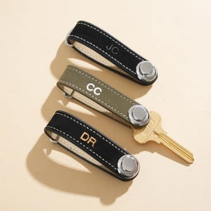 Può includere: Tre portachiavi personalizzati in pelle scamosciata nera e verde oliva con cuciture bianche. Ogni organizer ha una chiusura a bottone argentata e le iniziali: "JC", "CC" e "DR". Una chiave dorata è accanto agli organizer.