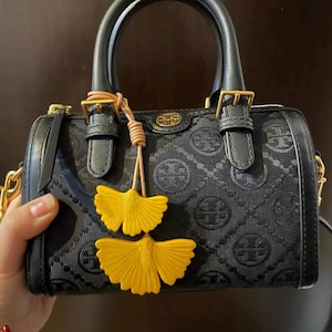 May include: Black handbag with a repeating pattern and gold-colored hardware. The bag has two handles and a decorative yellow leaf charm. The bag is a cylinder shape with a zipper closure.