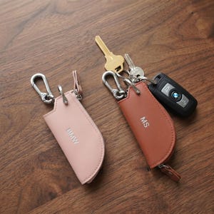 Puede incluir: Dos llaveros de cuero, uno rosa y otro marrón, con herrajes plateados y cremalleras. El llavero rosa tiene impreso "BMW", y el llavero marrón tiene impreso "MS". Las llaves y un mando de coche están adjuntos.
