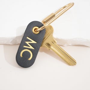 May include: A black leather key fob with gold-colored initials "MC" and a gold-colored key ring. A gold-colored key is attached to the fob. The key fob is a stylish accessory for organizing keys.