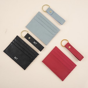 May include: A collection of leather cardholders and keychains in black, light blue, and red. Each cardholder has multiple slots and is paired with a matching keychain. The black cardholder and keychain are marked with the initials "MT", and the red cardholder is marked with "JAY".